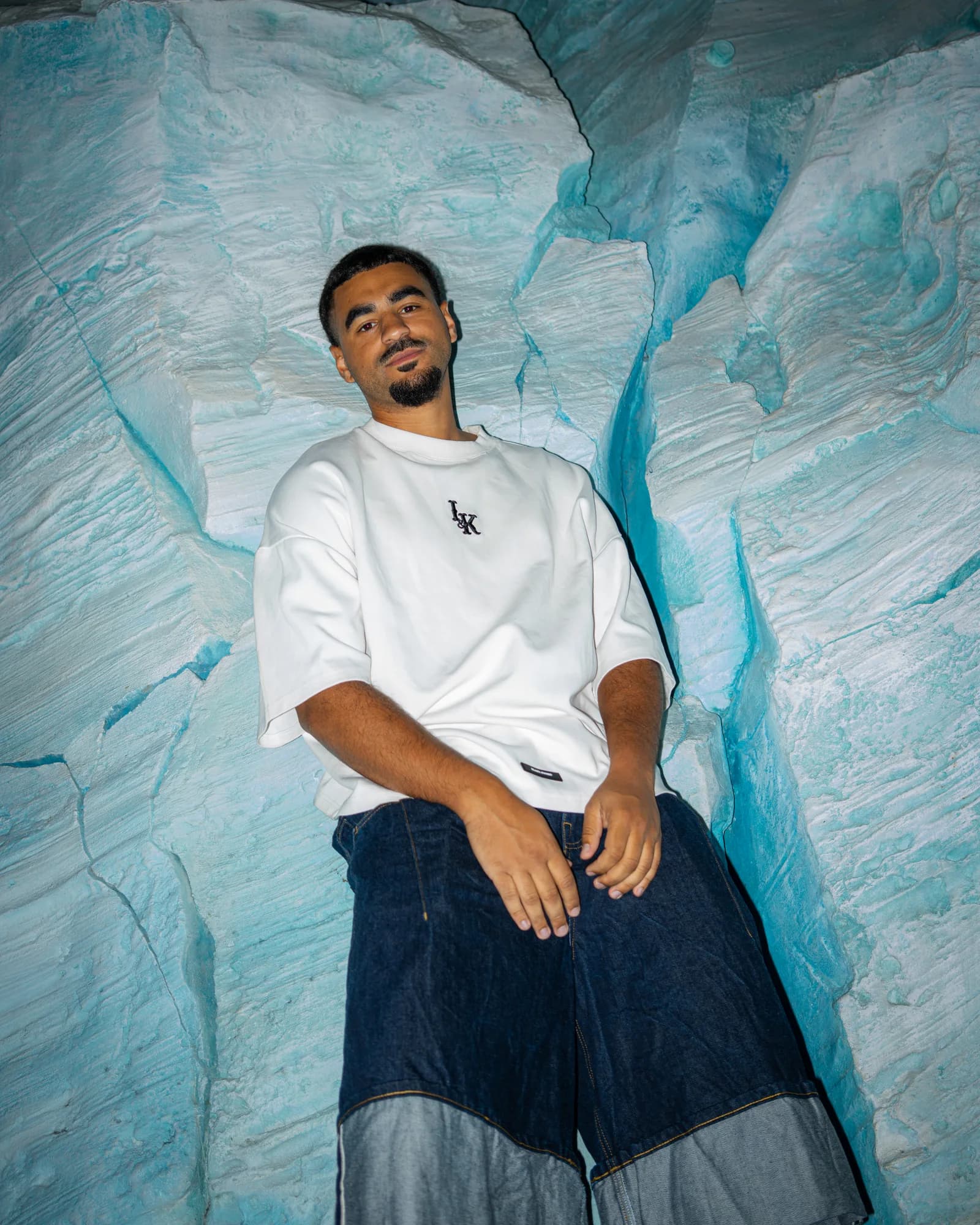 Barros leans into a turquoise ice-blue rock face, flash-lit in an oversized white tee and denim.