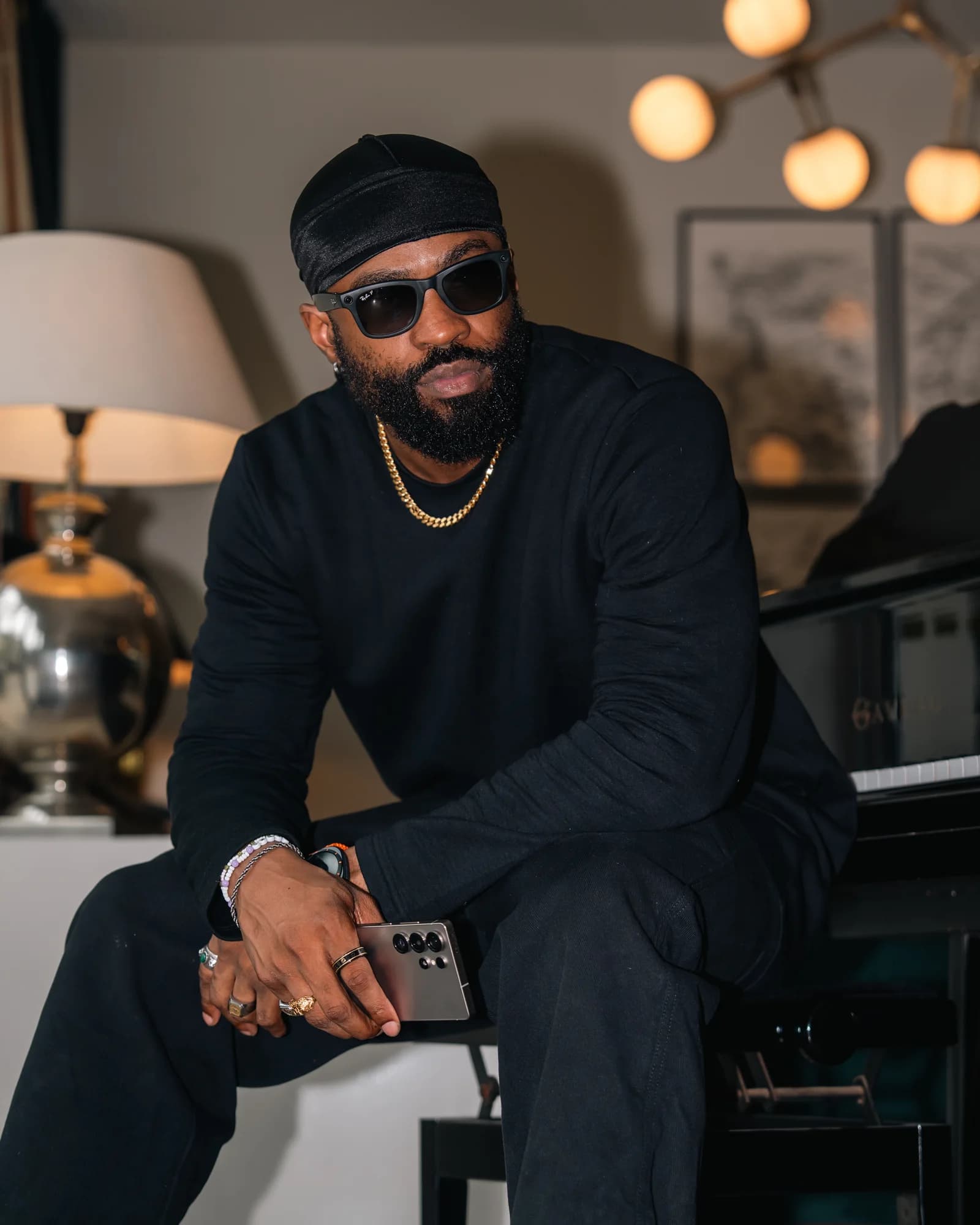 Brawks leans on a grand piano in shades and gold chain, phone in hand under soft ambient light