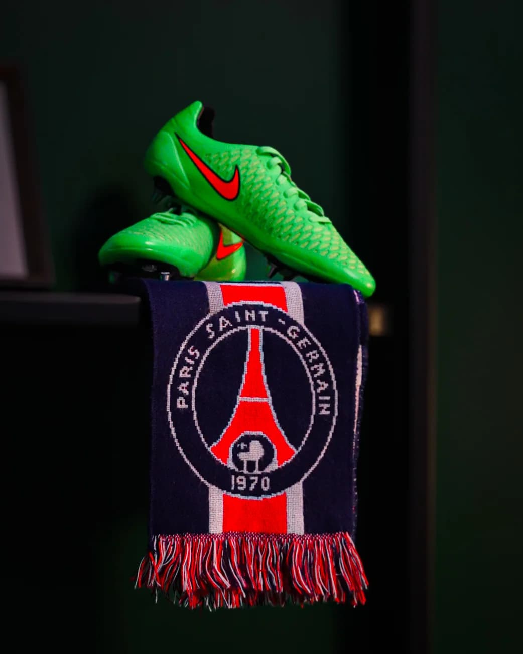 Green Nike boots resting on a draped Paris Saint-Germain scarf, a quiet tribute in the shadows.