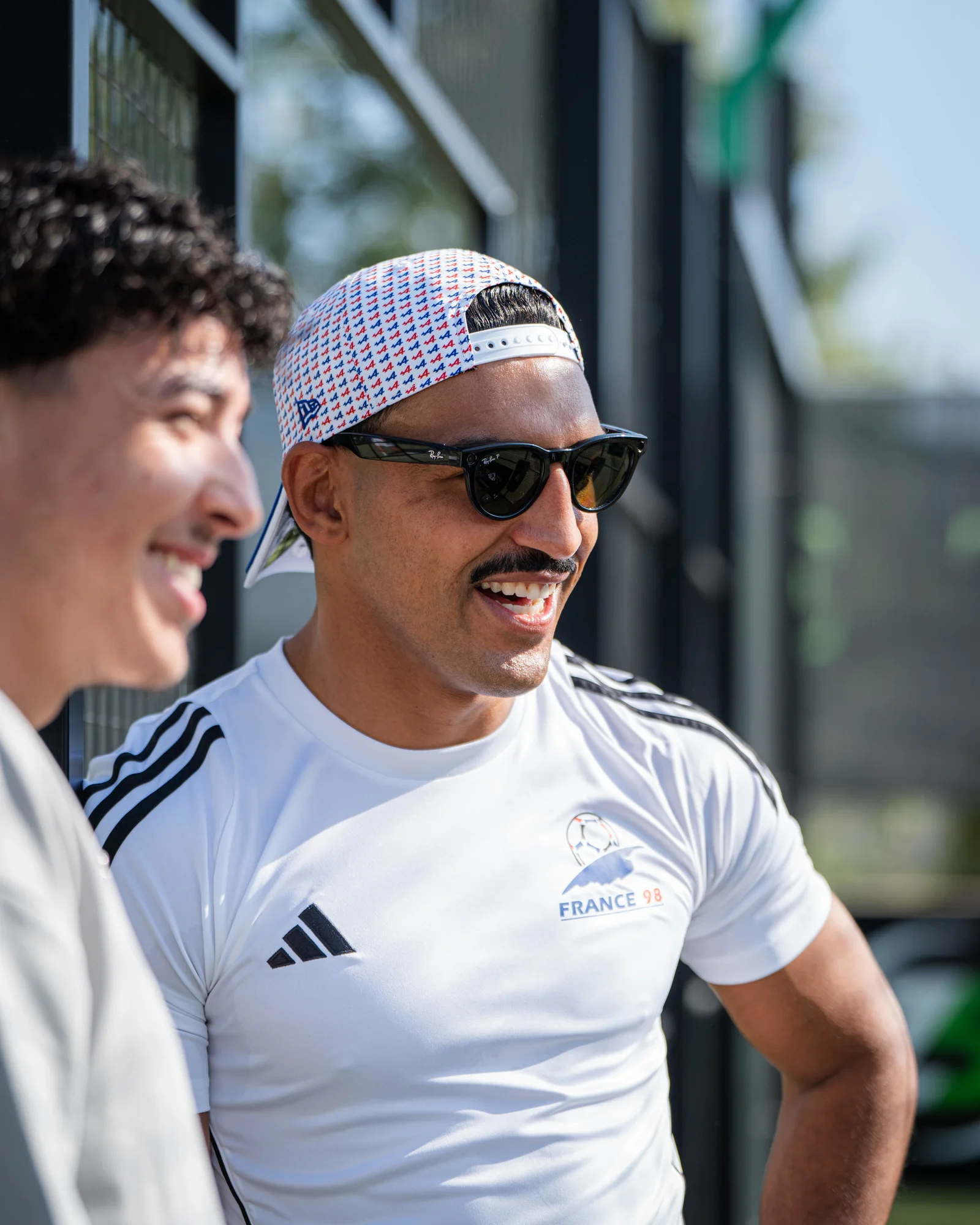 Candid close-up of a smiling player in Ray-Ban shades and a tricolor New Era cap, sharing a laugh courtside.