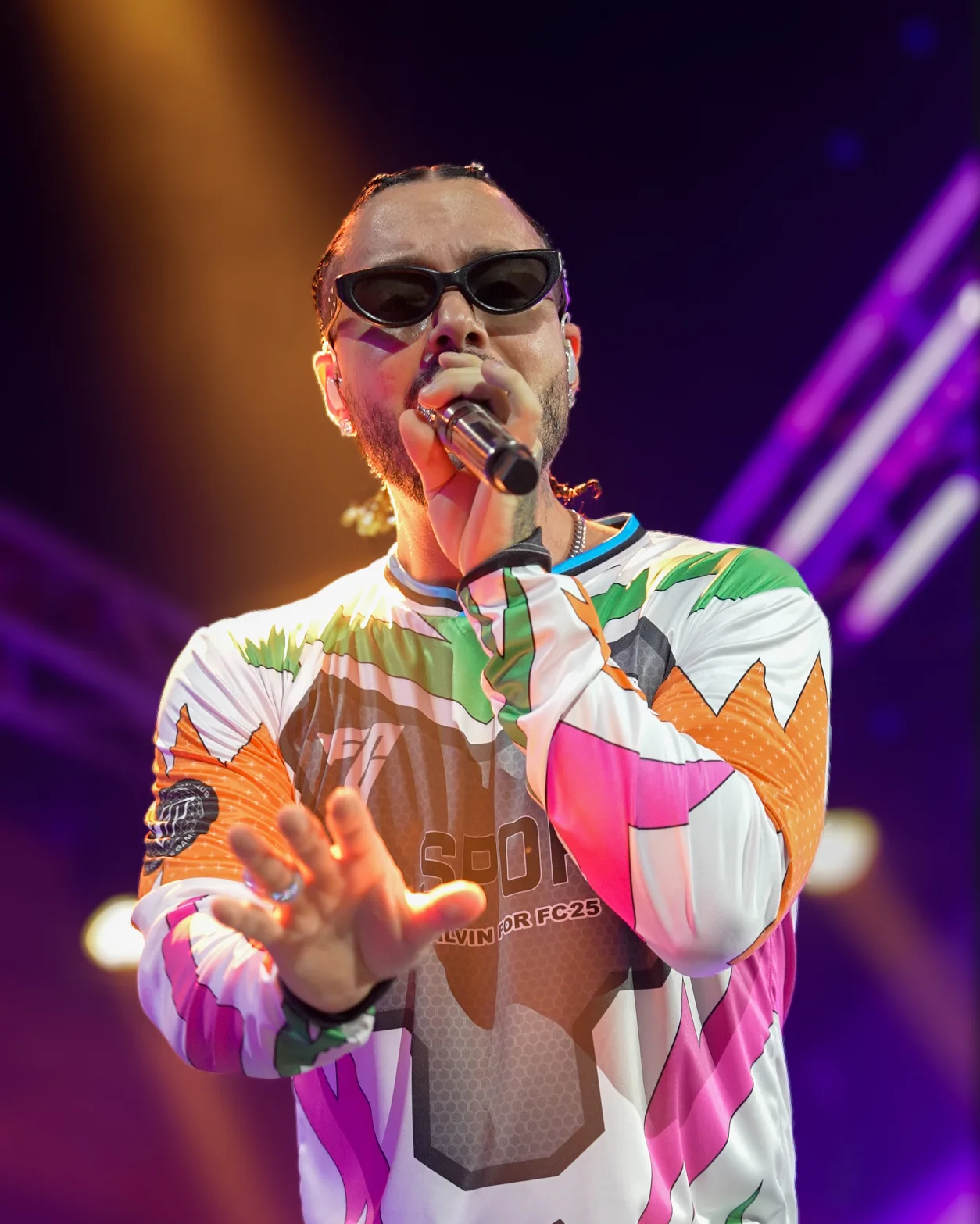 Low-angle portrait of J Balvin reaching toward the lens, his vivid EA Sports FC jersey glowing against purple beams.