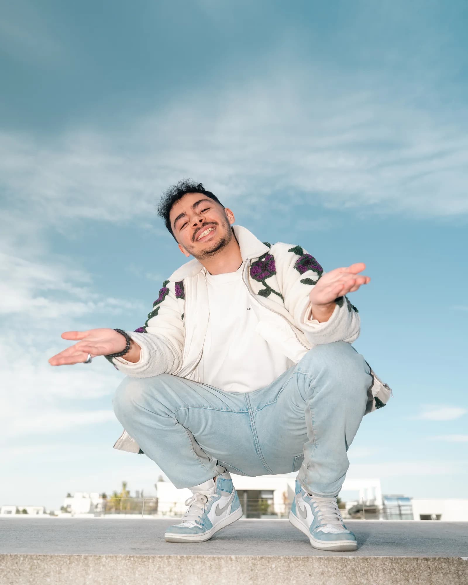 Playful low-angle portrait crouching on a ledge, arms open in a carefree shrug against a wide blue sky.