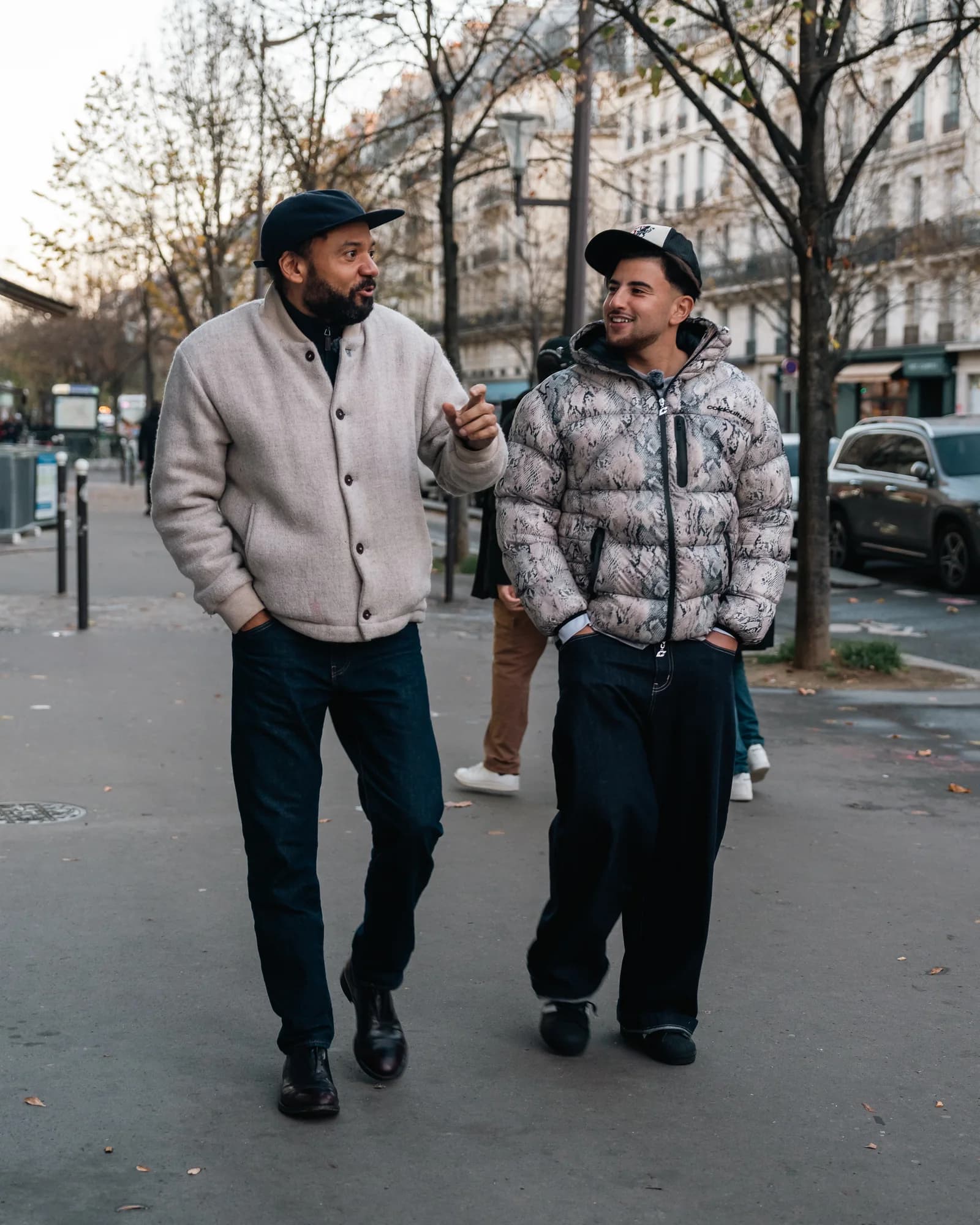 Riadh and Fabrice Éboué stroll side by side down a tree-lined Parisian boulevard, deep in conversation under the autumn light.